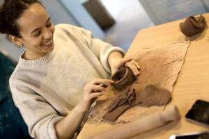 Photograph of Saskia Dakin working a small clay piece on a table, smiling, indoors.
