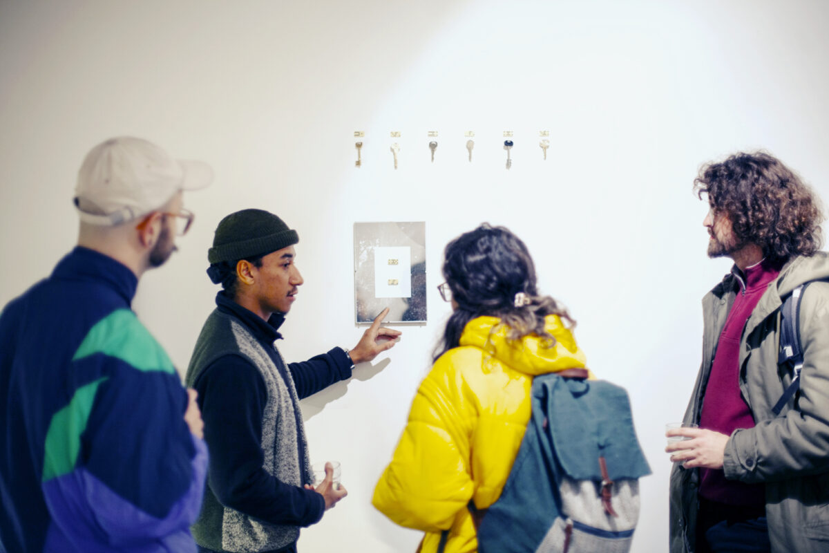 J D Rooney pointing at one of his works and speaking to visitors of his show, in a bright gallery space.