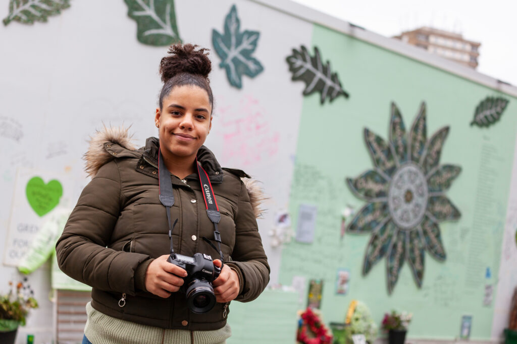 Ashleigh Ramel, previous Young Photographer, standing with a camera in front of the Grenfell flower and leaf mosaics.