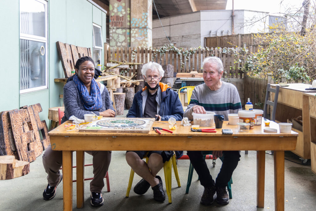 Three Maxilla Men's Shed participants sitting outside around a table with an unfinished mosaic
