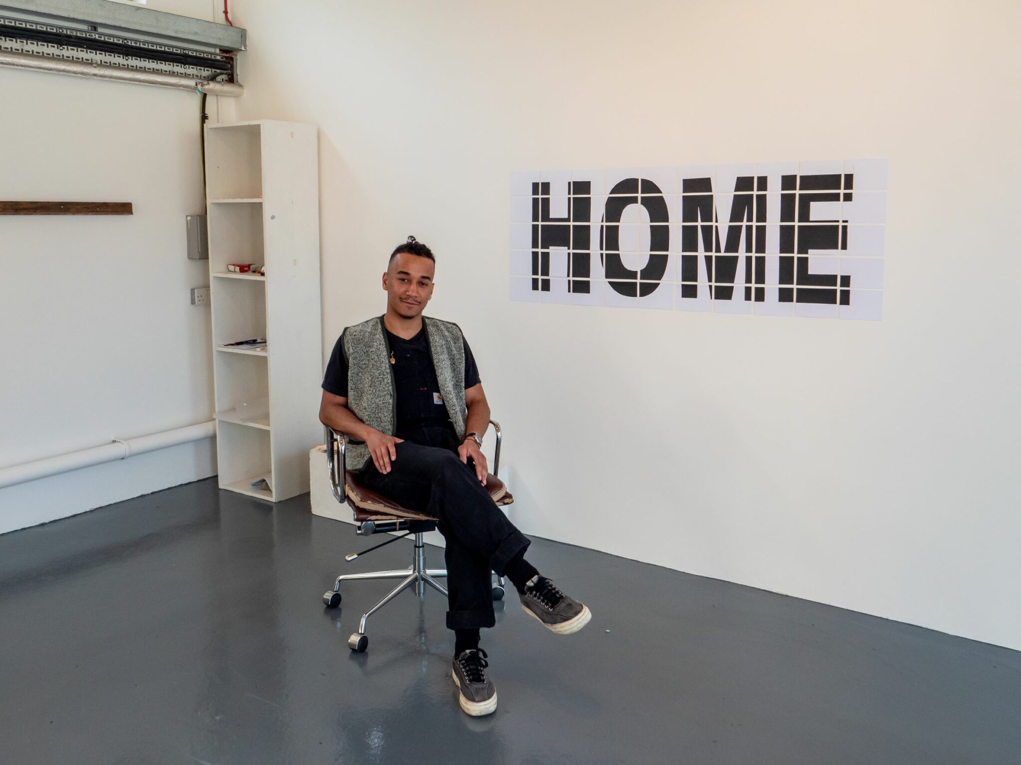 Programme participant sitting on a chair in their studio in front of a sign that reads 'HOME'