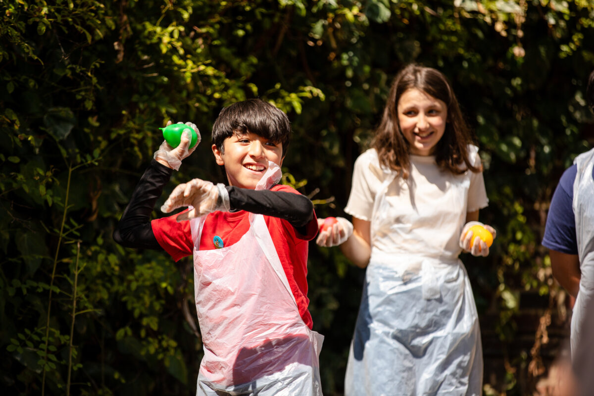 A group of children throwing paint balls and laughing, outdoors on a sunny day.