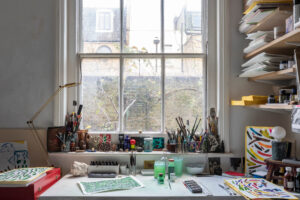 Closeup of a table by a window in a quiet artist's studio, the window overlooking onto a green residential street.