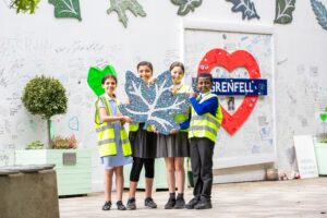Four children in school uniform pose holding a large mosaic leaf, behind them the hoarding at the base of Grenfell Tower.