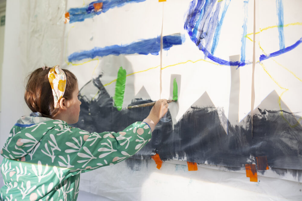 Child holding a paintbrush and painting a green stripe on a collaborative wall painting.