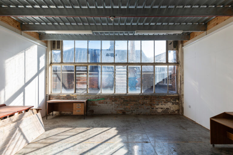 View of a bright but empty artists studio in an industrial building, a large window at the back.