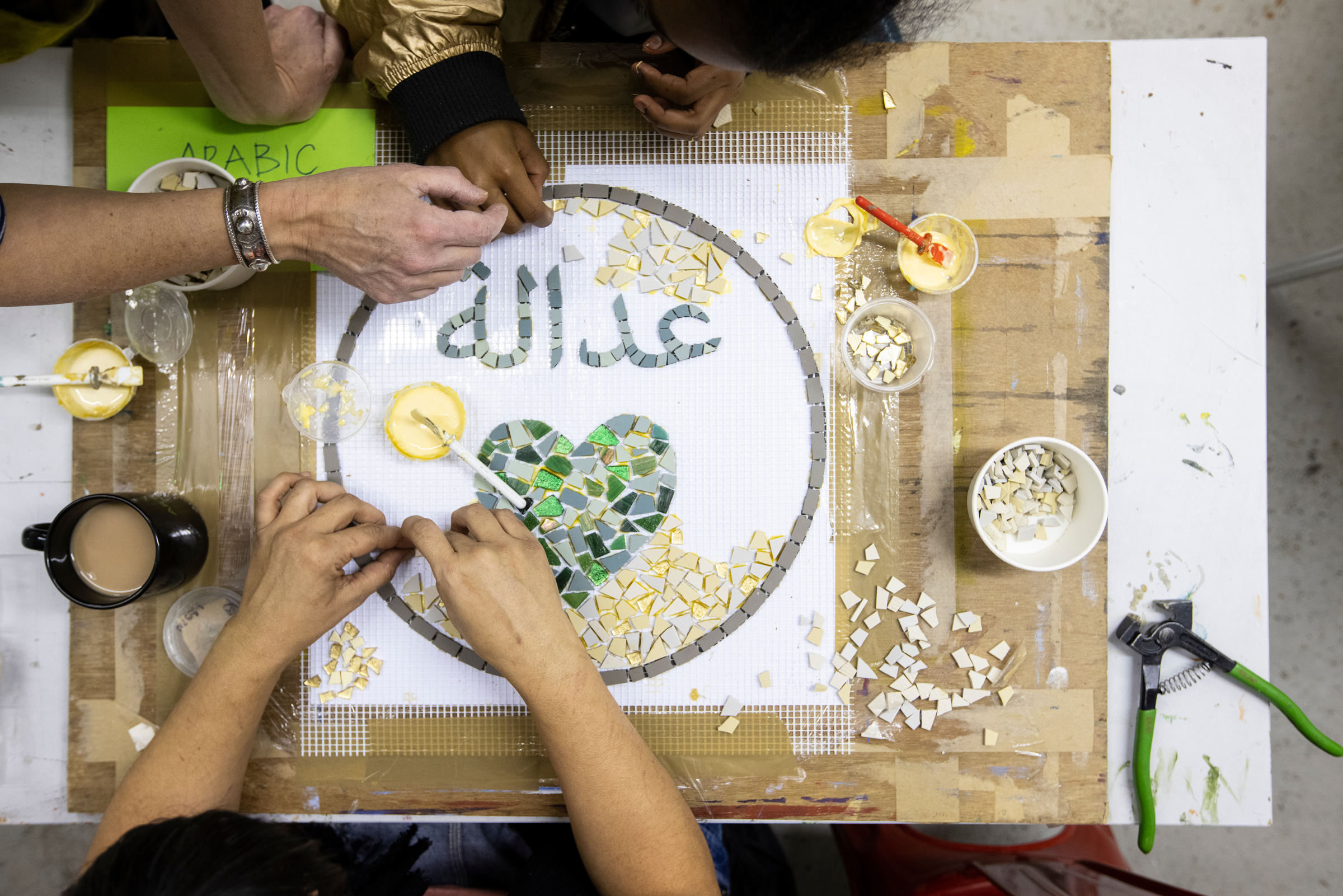 Birdseye view of various hands working together on a circular mosaic, on a table, indoors.