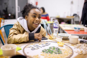 Closeup photograph of a child smiling across a table whilst working on a circular mosaic, indoors.