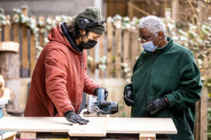 Photograph of two people using a circular saw on a workbench outdoors, a wooden fence behind them