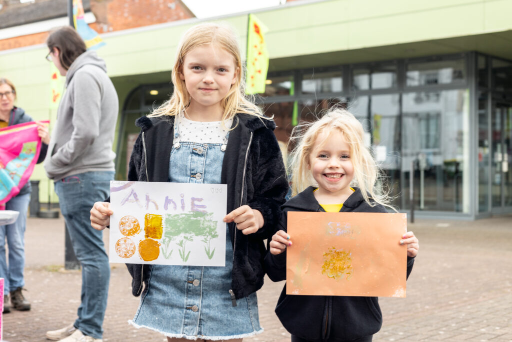Photograph of 2 children holding handmade prints and looking at the camera smiling, outdoors