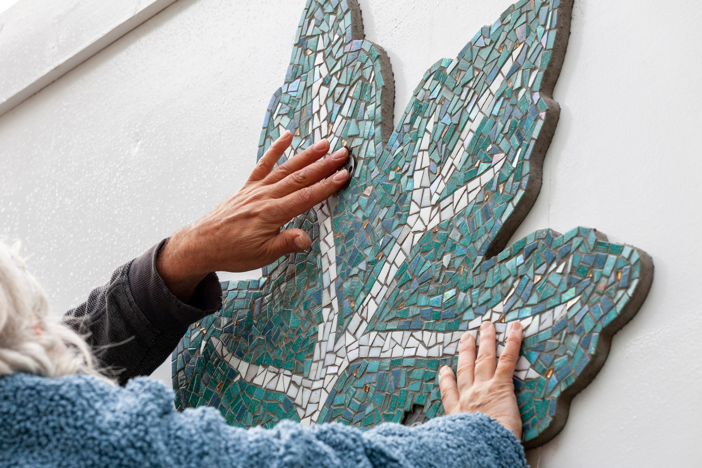 Closeup of two people's hands touching a leaf-shaped mosaic fixed to a white wall, outdoors.