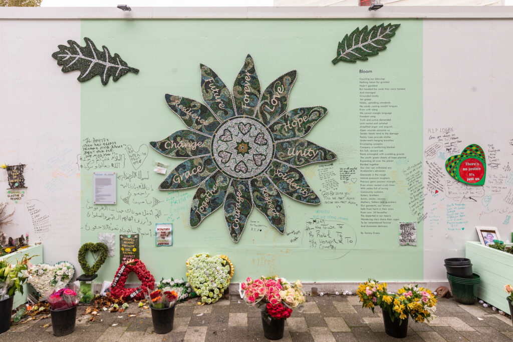 Flower-shaped mosaic in green and beige tones and leaf-shaped mosaic pieces installed on hoarding at the foot of Grenfell Tower, several bouquets of flowers against the hoarding, on the ground, hand-written messages on the wall.