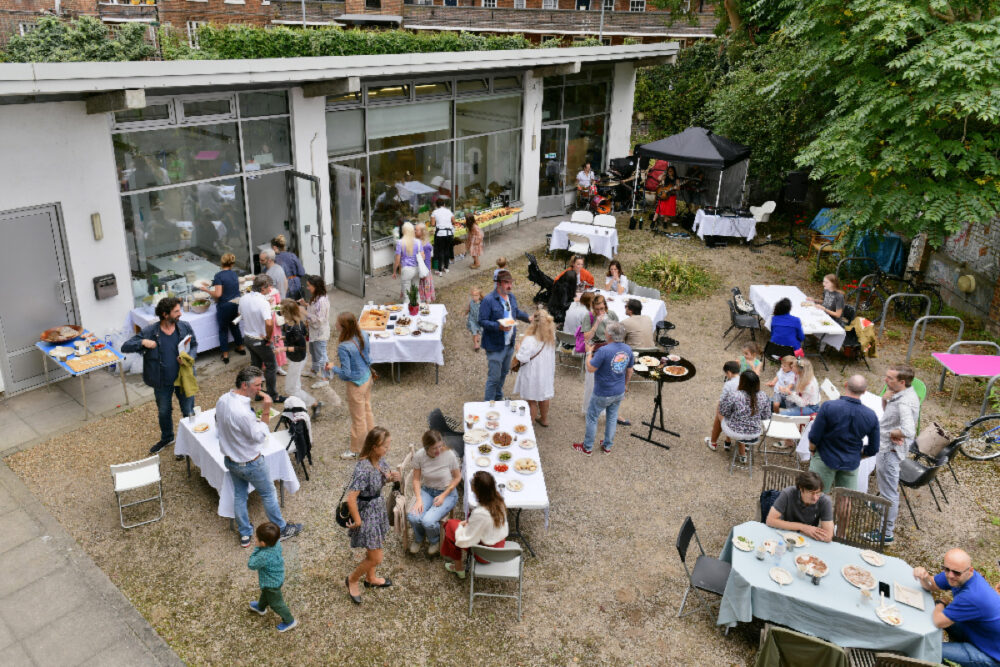 Photograph taken from above looking over an event outdoors, adults and children sitting and standing around tables with food, and live music.