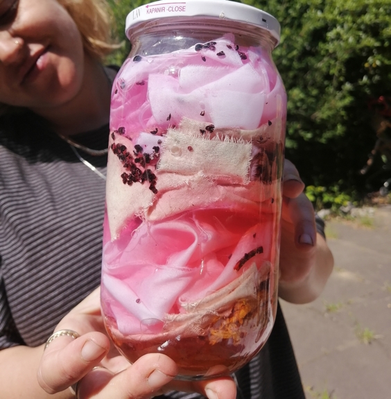 Closeup of a glass jar containing naturally dyed pieces of pink cloth, held by a person, outdoors in a green space.