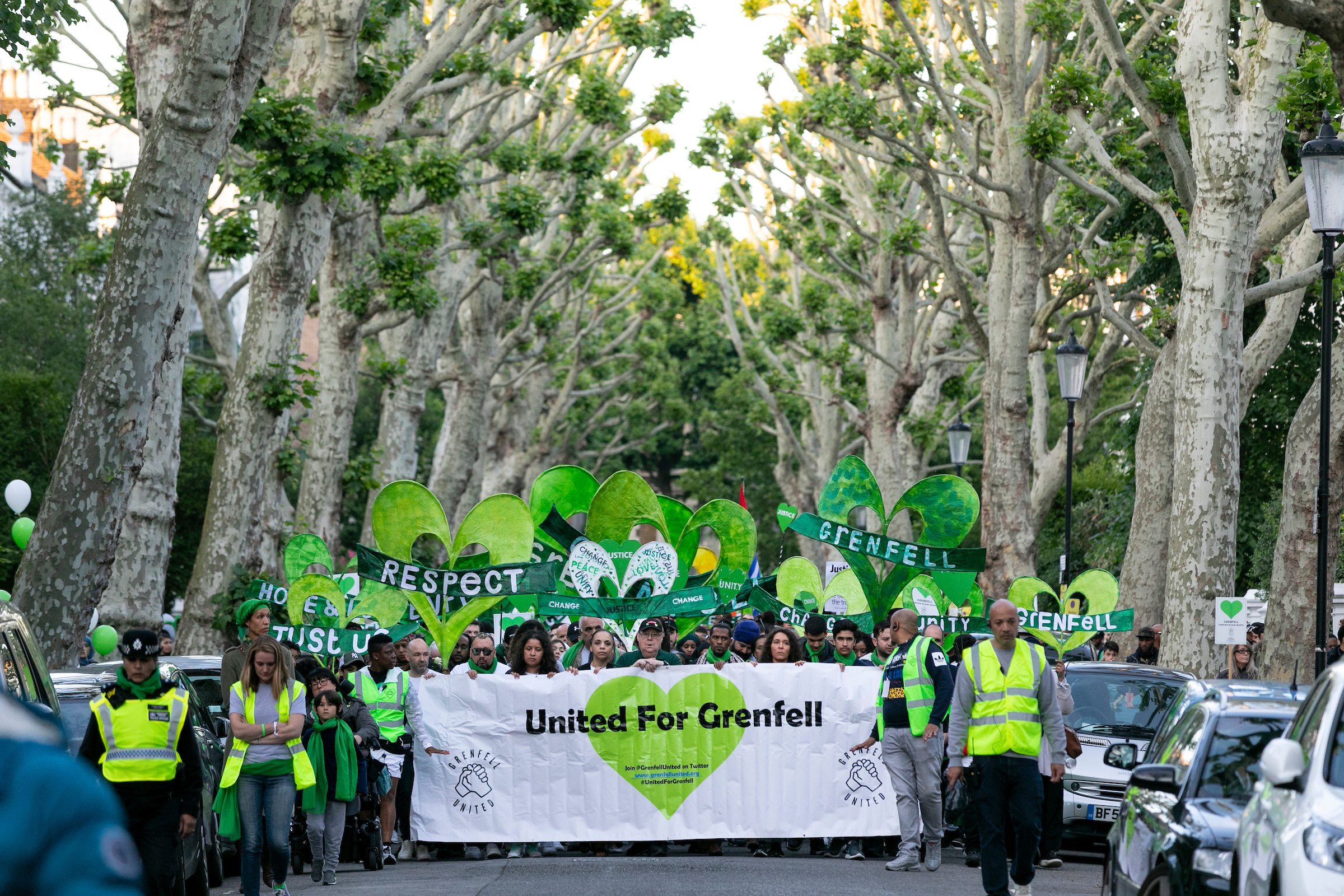 Photograph of a group of people walking peacefully, holding green hearts and banners, on a road flanked by large tall trees