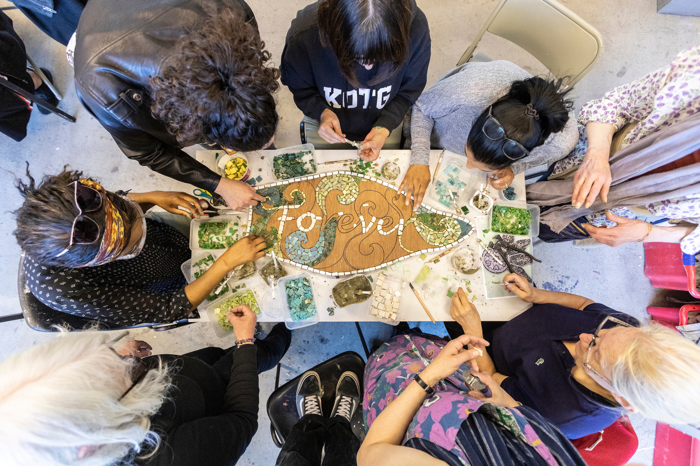 Bird's eye view of a group of people working together on a petal-shaped mosaic artwork, around a table, indoors