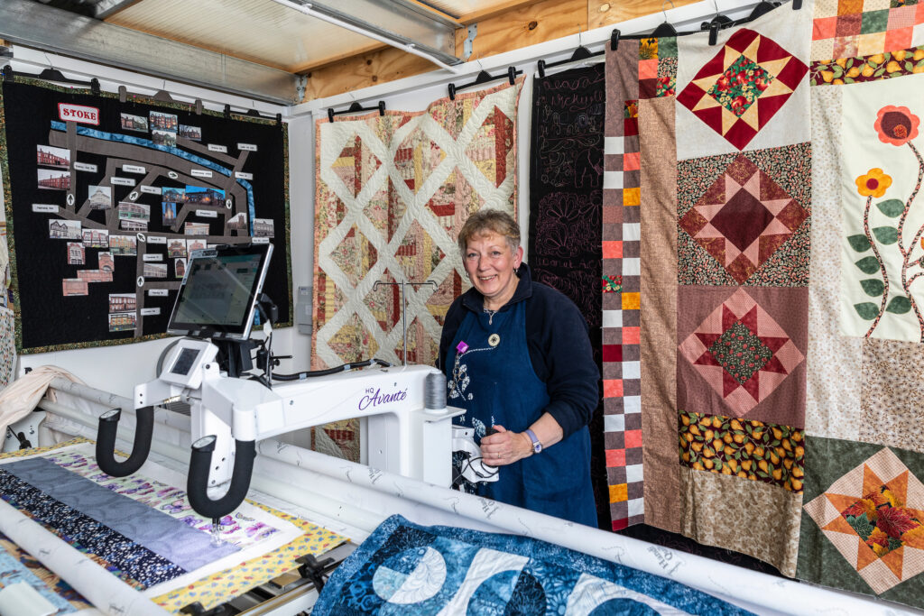 Photograph of Nicky Griffiths operating a quilting machine in her studio, finished quilts in earthy colours hanging behind her.
