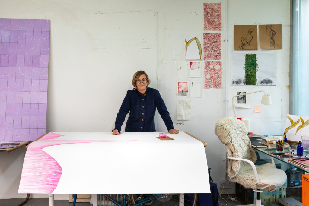 Photograph of artist Lisa Dredge posing hands on table looking at the camera, a large drawing on paper falls over the sides of the table, in an artists studio, indoors.