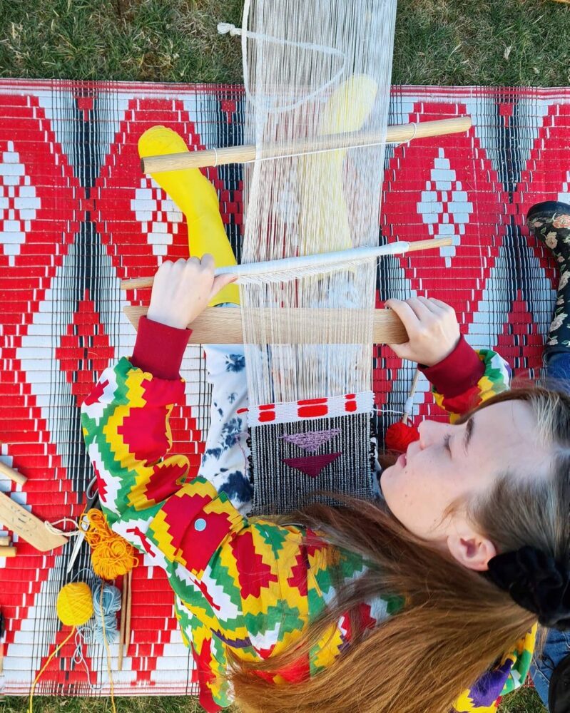 Close up of a person using a backstrap loom to work on an orange and yellow tapestry piece, indoors
