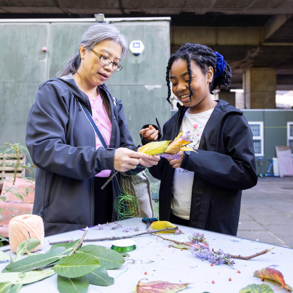 An adult and a child playing together to build a magic wand out of natural forest materials, standing around a table, outdoors in a creative setting.