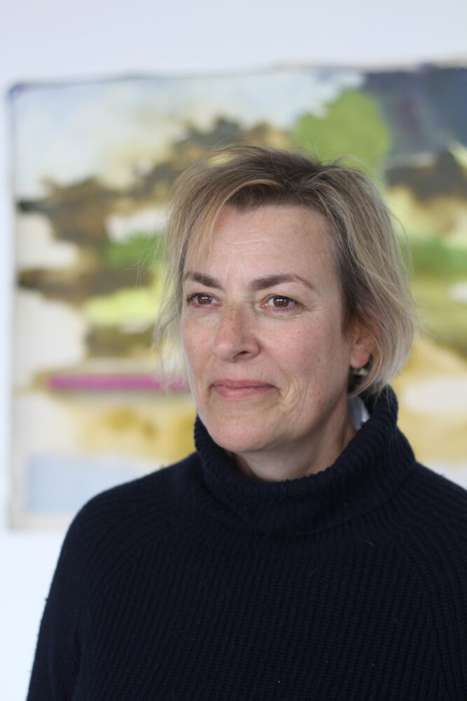 Portrait of Amanda Jackson wearing a black turtle neck, one of her artworks blurred in the background.