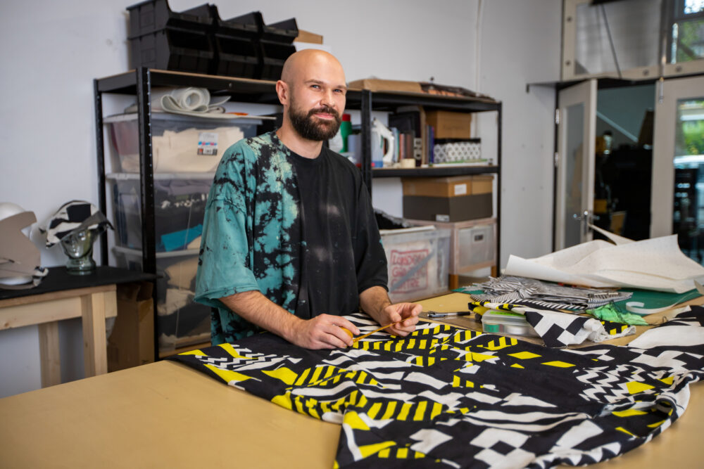 Photograph of Douglas Atkinson creating clothes in an artist's studio, sitting and smiling at the camera.