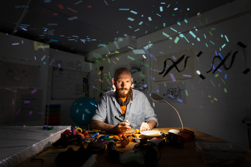 Artist Ian Gouldstone working on small electronics on a table in his studio, lights off, his face illuminated by a table lamp and, behind him, colourful geometric light forms move around the walls illuminating the darkened studio.