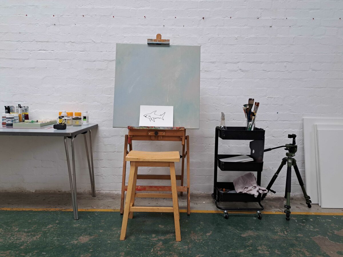 Image description: Photograph of an easel with a medium-sized monochrome painting and an A4 photocopy depicting a shark cartoon leaning against it, in an artist's studio.