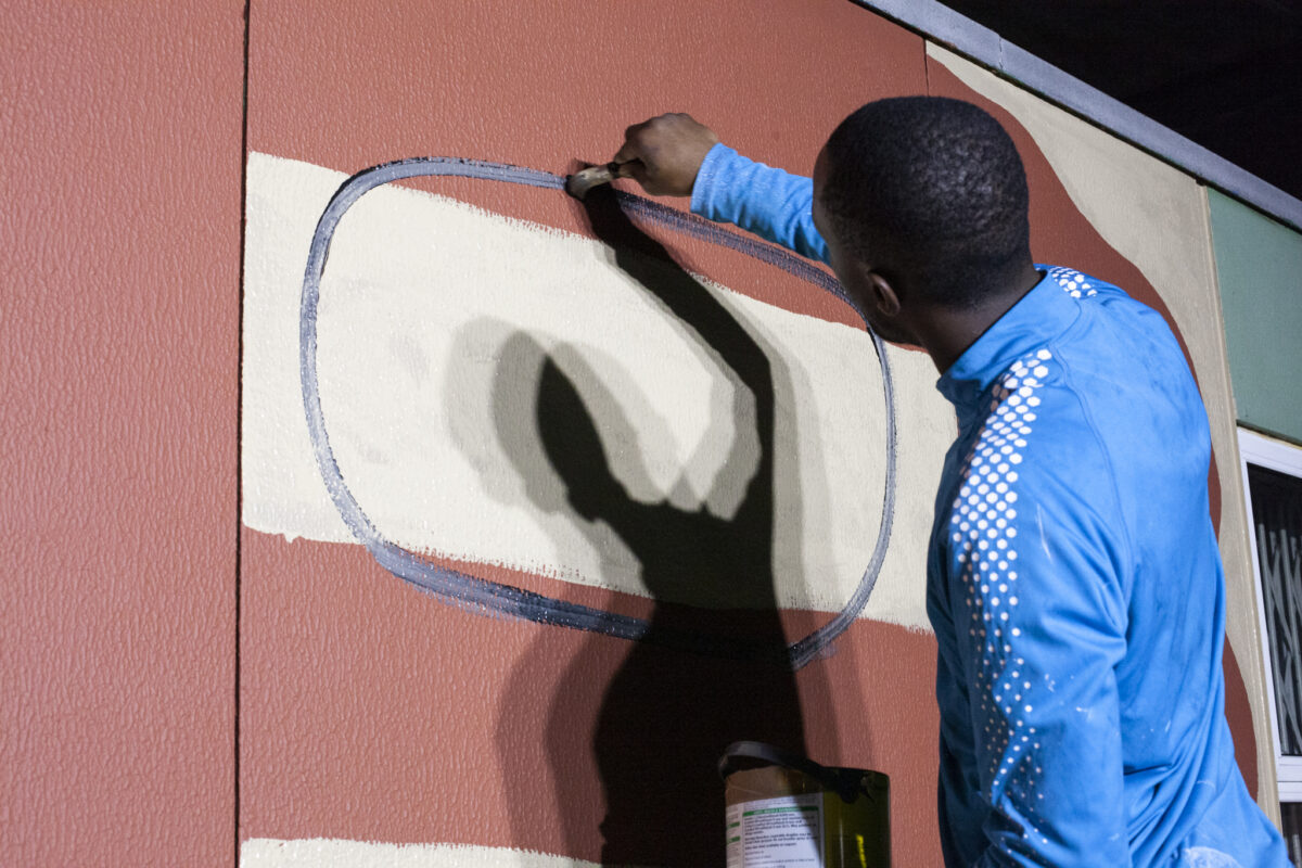 Photograph of artist Laho Jebak using a brush to create a large painting in the Shed's outdoor space, in the evening.