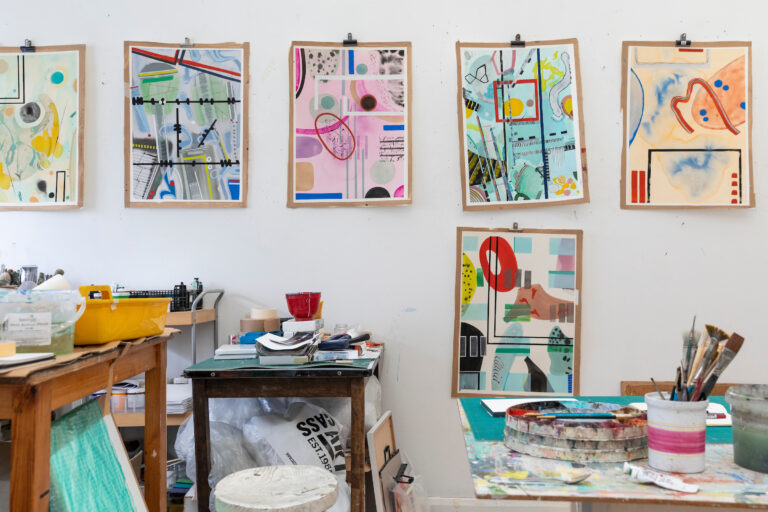 Artist's studio with colourful abstract watercolours on the wall, art materials in use on small tables.
