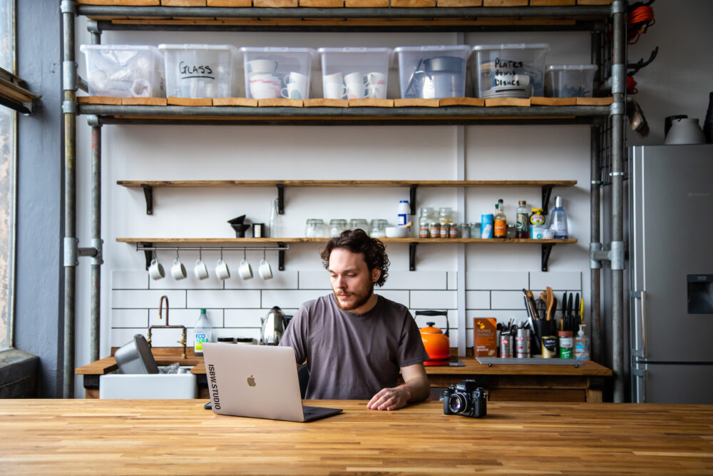 Photograph of a creative person working on a laptop, behind a large wooden table in a bright artists' studio.