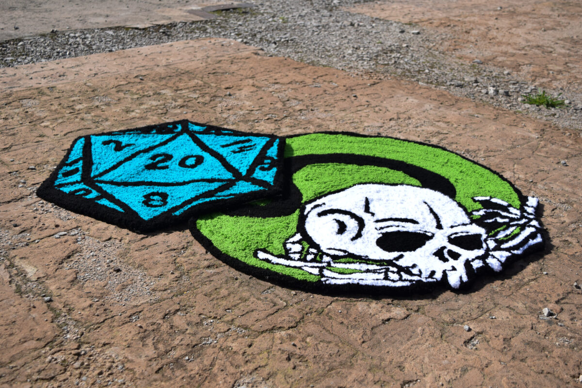 Rug depicting a polyhedral dice and a skull on the ground, outdoors.