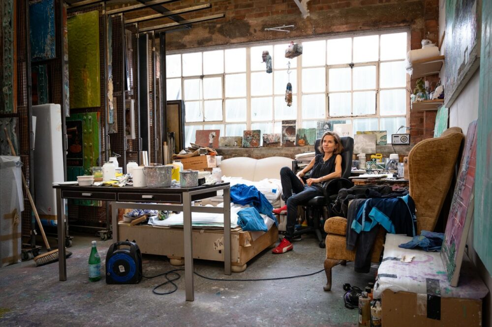 Miranda Donovan sitting on a lounge chair in a spacious artist's studio, large paintings stored vertically on the left, a large window at the back.