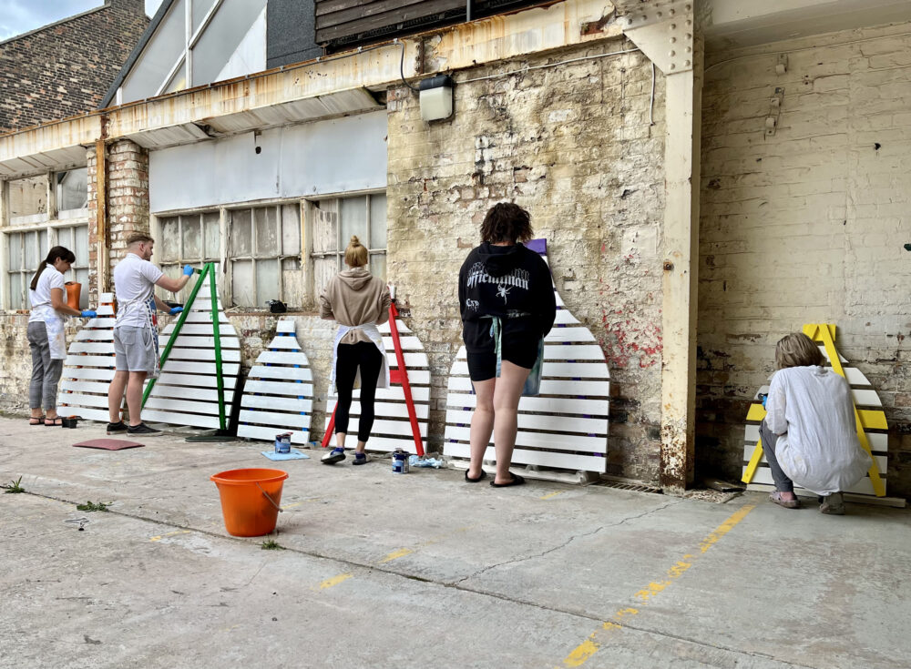 A group of people painting a kiln-shaped wooden trellis, outdoors, against a building wall.