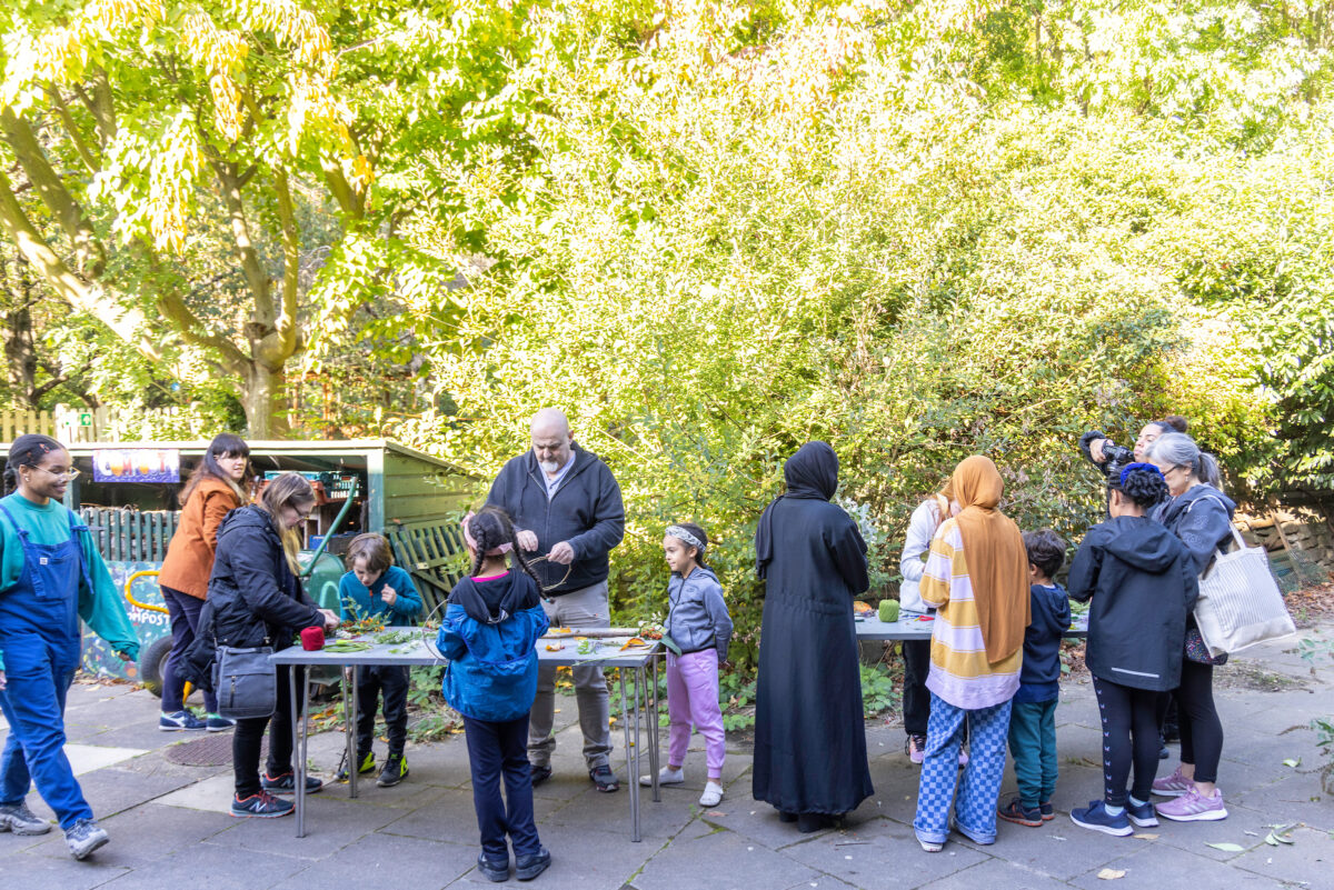 A group of families creating magic wands from natural objects found in the forest garden, around large tables, outdoors in a park.