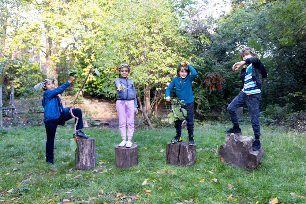 Four children standing on tree trunk stubs wearing handmade costumes with materials from the forest, smiling and posing for the camera, outdoors in a park.