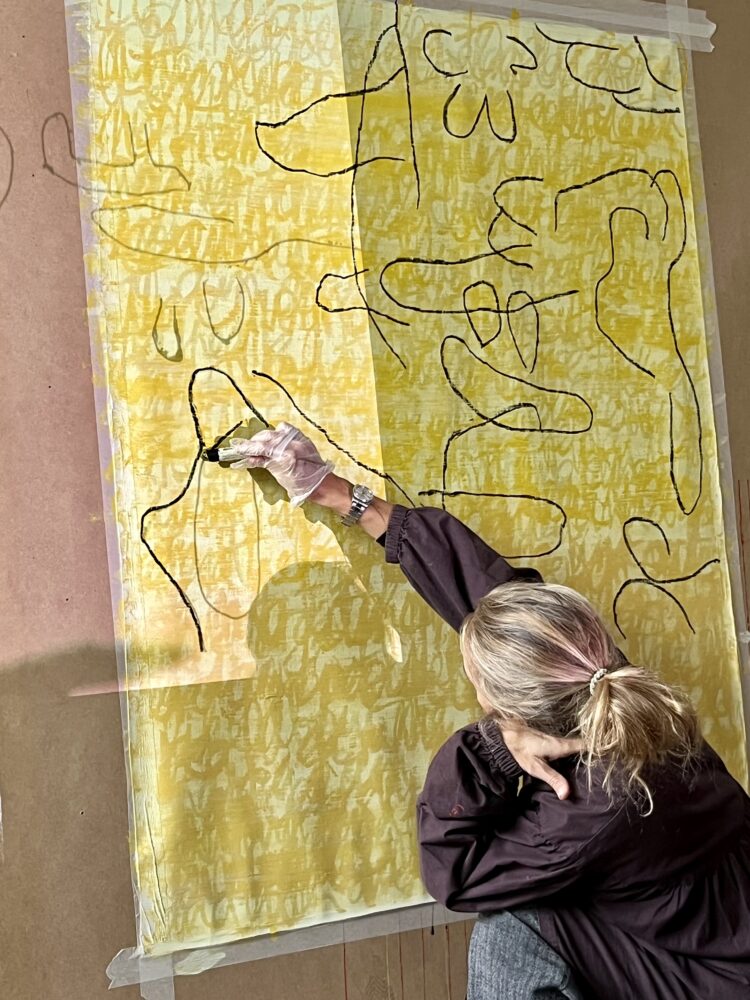 Closeup photograph of Florence drawing black lines on a yellow painted canvas taped to a board, using a black oil pastel crayon.