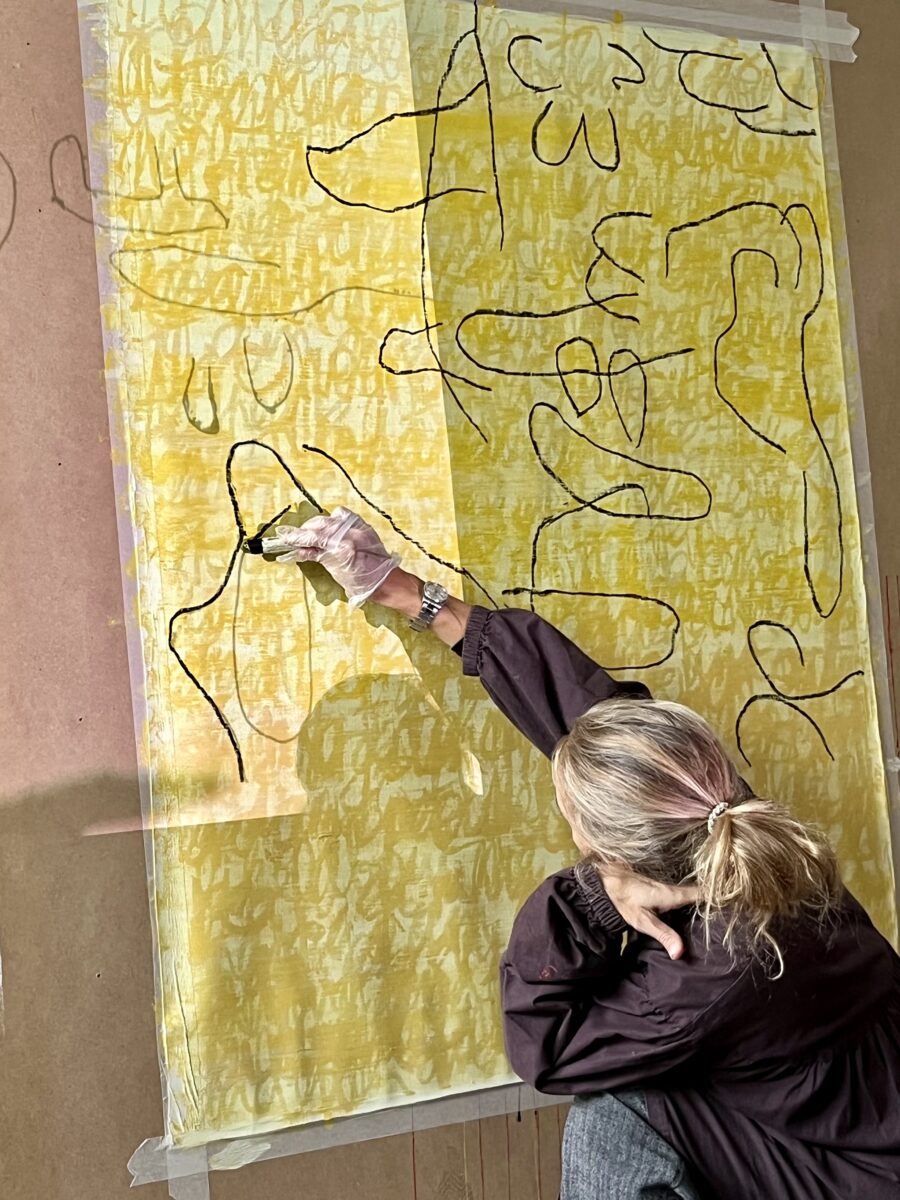 Closeup photograph of Florence drawing black lines on a yellow painted canvas taped to a board, using a black oil pastel crayon.