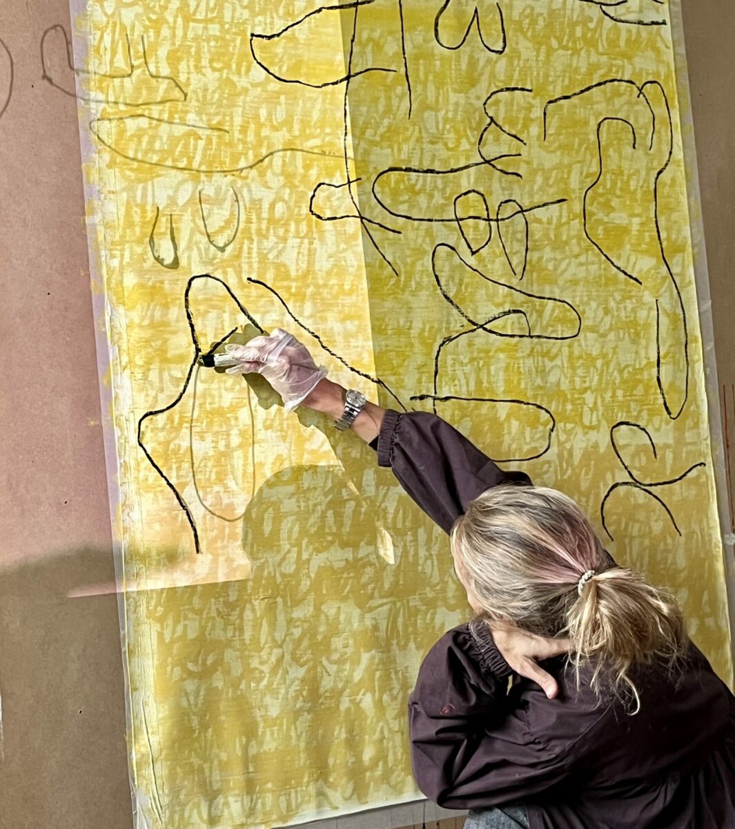 Closeup photograph of Florence drawing black lines on a yellow painted canvas taped to a board, using a black oil pastel crayon.