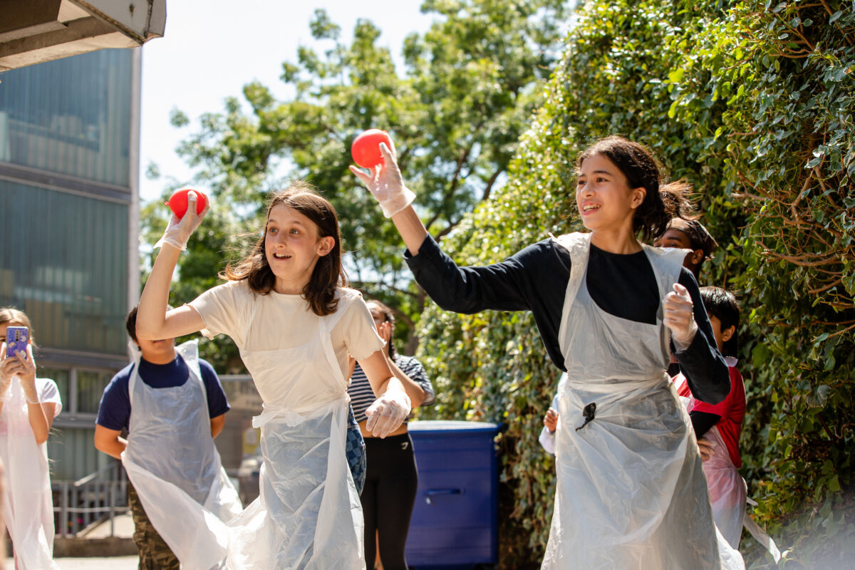 A group of children throwing paint balls and laughing, outdoors on a sunny day.