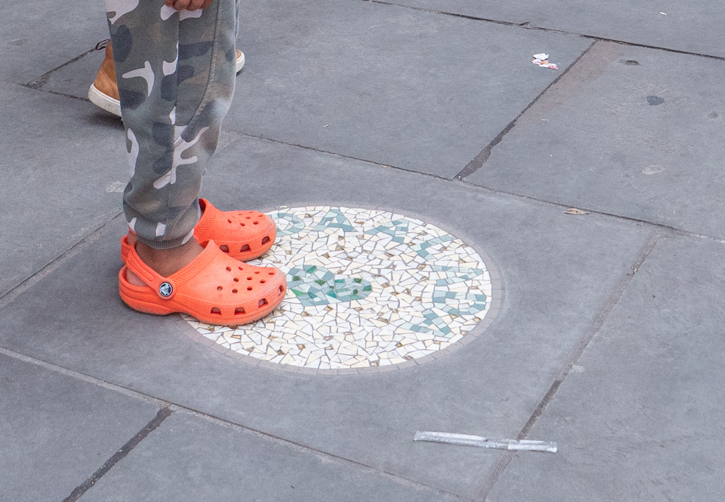 Closeup of a child's feet wearing orange crocs stepping onto a circular mosaic artwork on a stone slab on the street, outdoors.