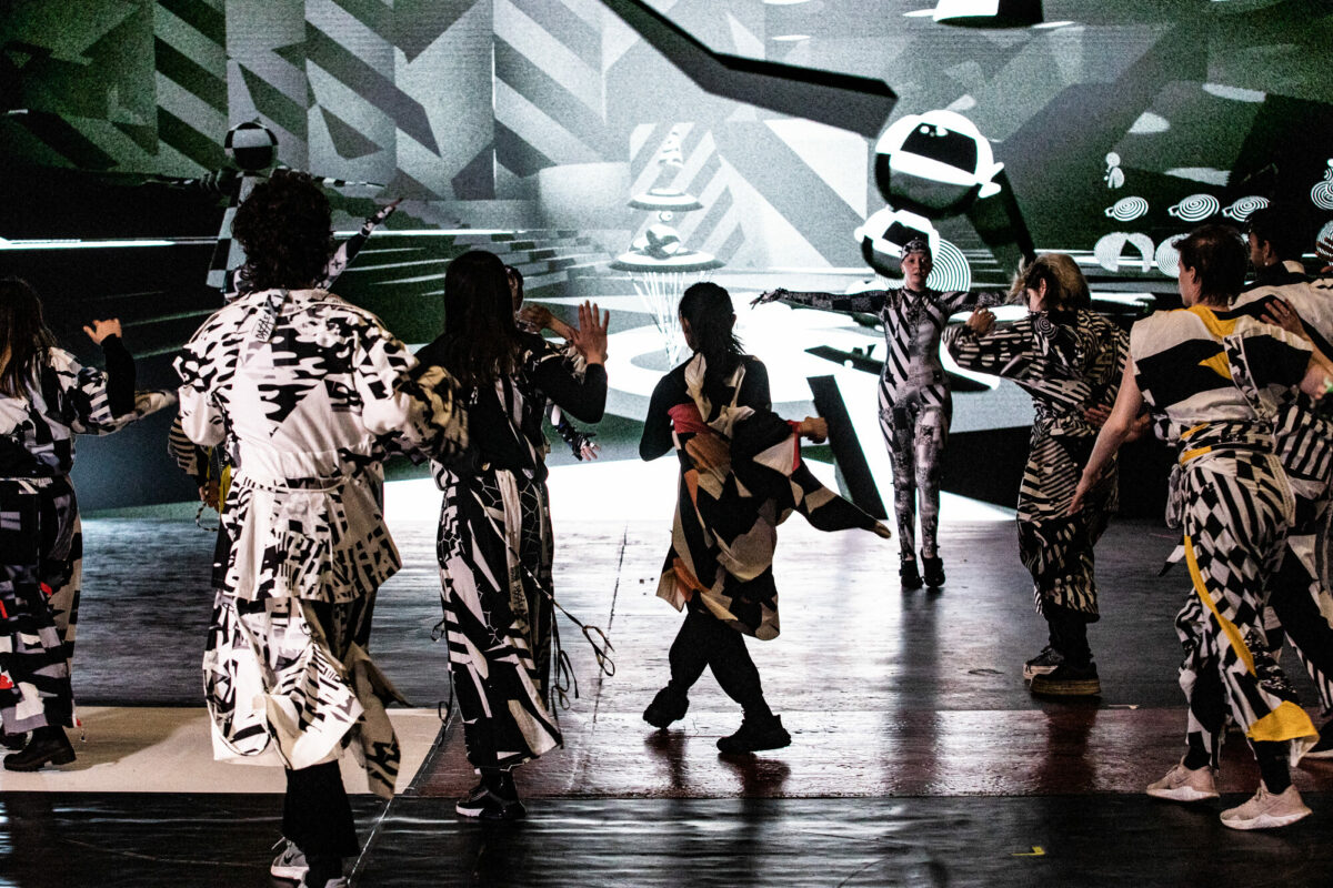 Photograph of a group of performers moving on stage, wearing contrasting black and white geometric patterned clothes, against a monochromatic backdrop in the same style