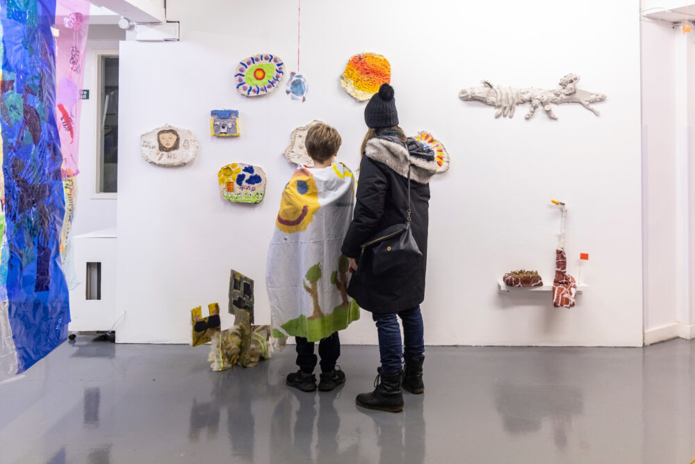 Photo of an adult and a child looking at wall-based artworks in a show, in a bright gallery space.
