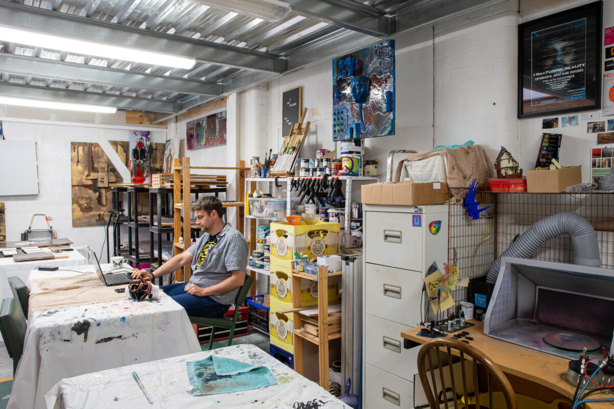 Photograph of an artist working at a desk in their studio, shelves and cabinets stocked with utensils and art materials.