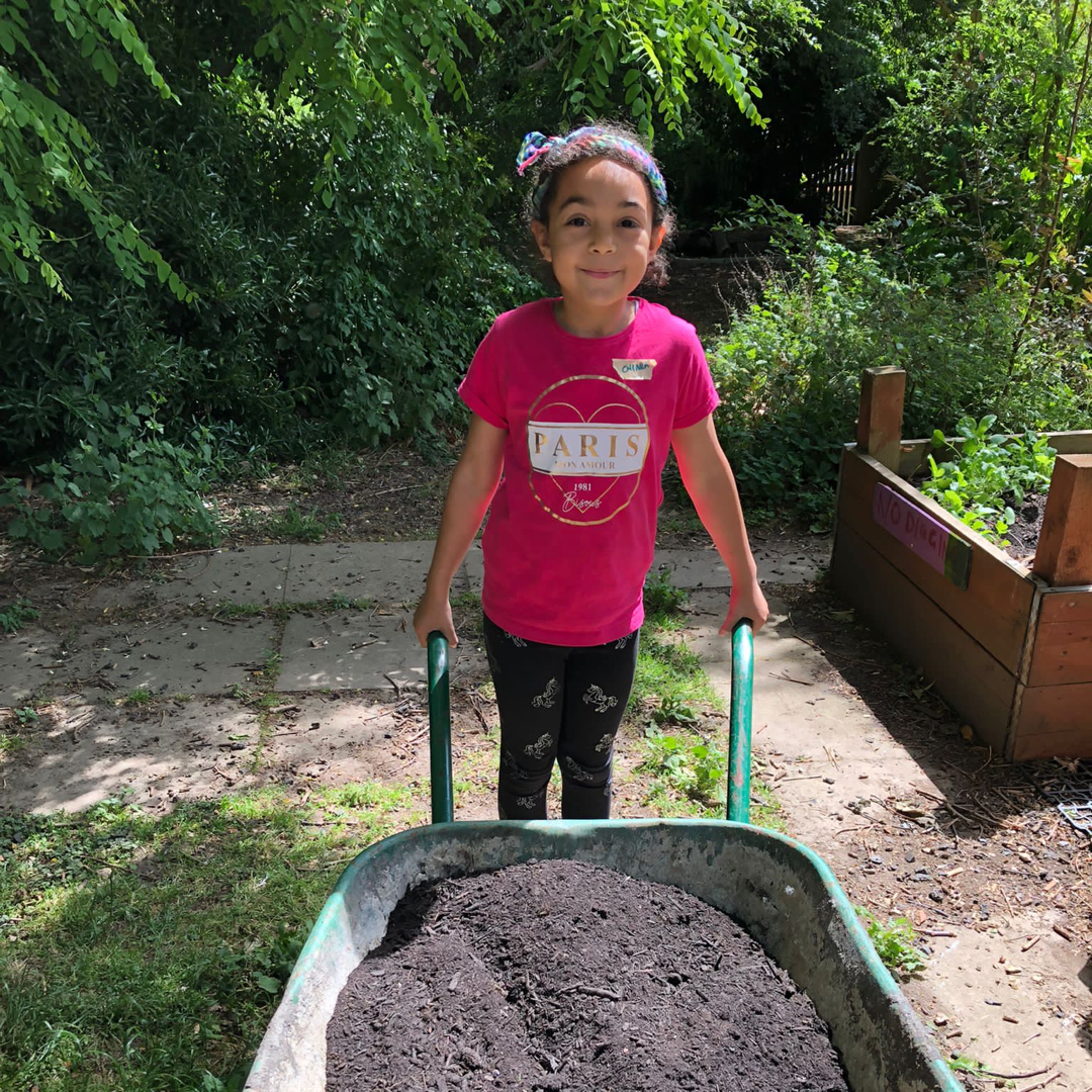 Photograph of a child holding a wheel barrow full of compost and looking at the camera, smiling.