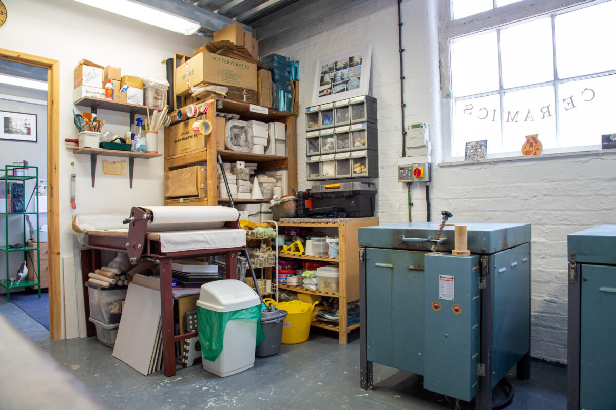 Photograph taken inside a professional ceramics studio with kilns, a presser, materials stacked on shelves.