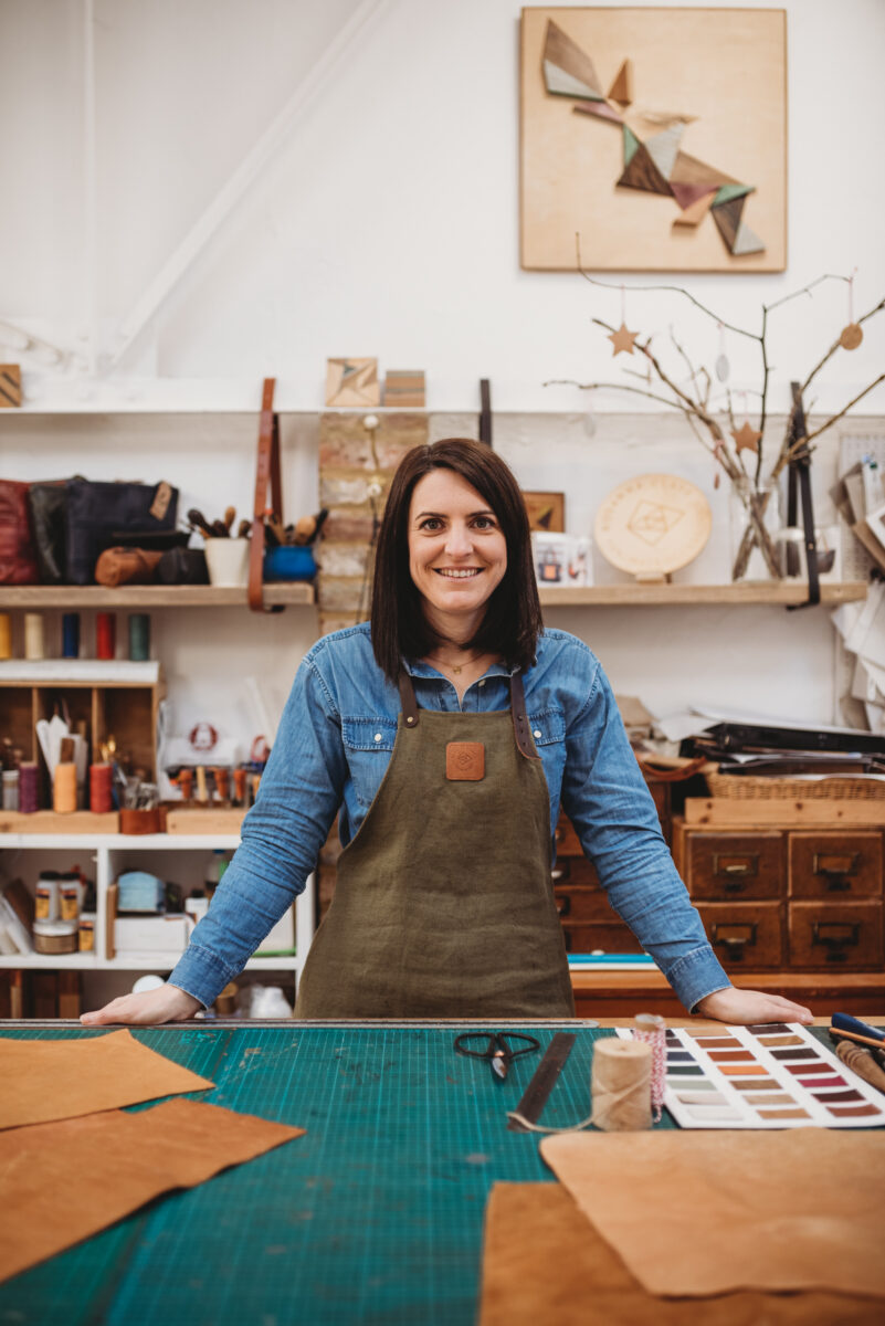 Photograph of artist Rosanna Clare in her leatherwork studio, hands on the workbench and smiling at the camera.