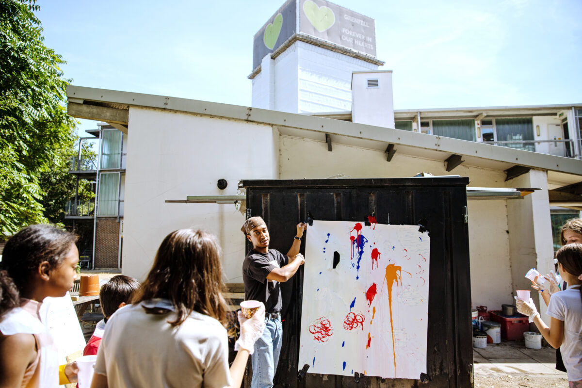 Artist Jack Rooney holding up a large sheet of paper with paint splatters on and speaking to a group of children, outside of ACAVA Blechynden Street Studios with the Grenfell Tower in the background.