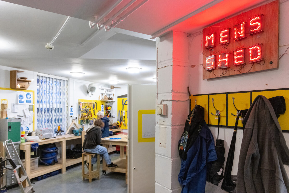 Entrance to Maxilla Men's Shed, a red LED sign above a coat hanger, programme participants in the background working together on workbenches, carpentry tools and materials on shelves and walls.