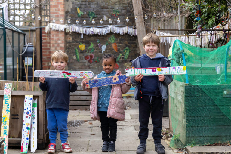 Photograph of three children posing for a photograph holding hand-painted wooden planks, next to raised garden beds in an urban garden, outdoors.